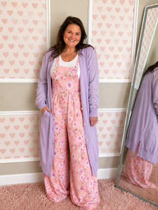 Woman wearing a pink floral jumpsuit with a purple cardigan in a room with heart-patterned wallpaper.