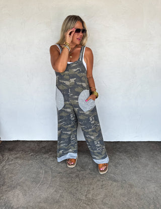 Front-facing view of a sleeveless camo jumpsuit featuring contrast trim, side pockets, and a relaxed wide-leg fit, styled casually with a white tank and sandals.
