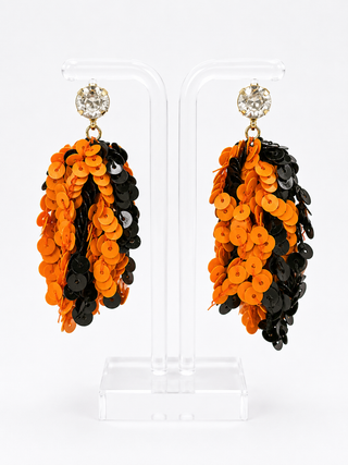 A pair of statement dangle earrings displayed on clear acrylic stands against a white background. The earrings feature a gold-toned setting with a clear rhinestone stud, adorned with cascading layers of bright orange and black sequins.