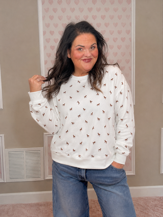 Woman wearing a soft white sweatshirt with mini embroidered brown horses paired with relaxed-fit denim jeans and western-inspired styling indoors.