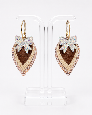 A pair of dangling earrings displayed on a clear acrylic stand against a white background. Each earring has a gold-tone hoop and a teardrop-shaped charm made from layered materials: a leopard-print outer layer, a textured brown center, and a glittery silver bow at the top.
