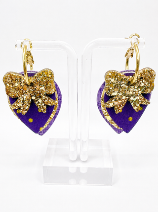 A pair of gold hoop earrings with purple and gold dangling charms shaped like a teardrop with a glittery gold bow on top, displayed on clear acrylic stands against a white background.