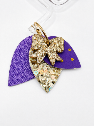 Earrings featuring three layered teardrop-shaped pieces: a textured purple piece on the left, a gold glitter bow on top, and a gold sequined purple piece on the right, attached to a clear hanger on a white background.