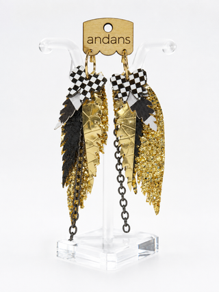 A pair of earrings gold-toned statement with black checkered accents, textured feathers, and glittery gold leaf details, displayed on a clear acrylic stand with a branded cardboard header.