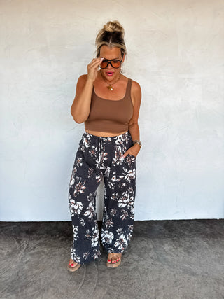 Woman wearing a brown tank top and floral pants against a white wall.