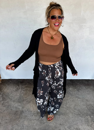 Woman wearing a brown top and black floral pants with a black cardigan against a white wall.