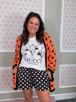 A woman modeling a cozy orange fuzzy cardigan with black polka dots, paired with a white graphic t-shirt and black-and-white polka dot shorts, standing against a wall with pink heart wallpaper.