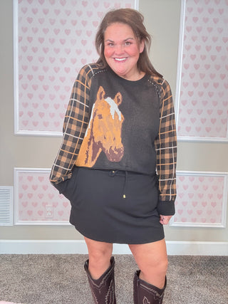 Woman wearing a dark black sweatshirt with a horse graphic, plaid shirt, and black skirt in a room with heart-patterned wallpaper.