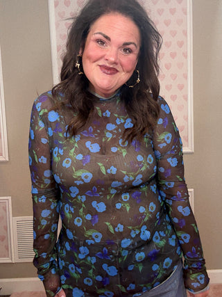 Woman wearing a sheer floral mesh top with long sleeves with a pattern of blue flowers on a brown background.