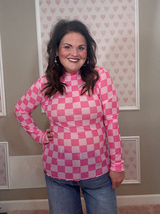 Person wearing a pink checkered mesh long sleeve top  standing in front of a wall with heart patterns.