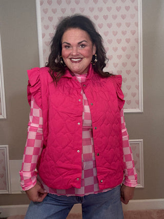 Woman wearing a pink quilted vest over a plaid shirt in a room with heart-patterned wallpaper.