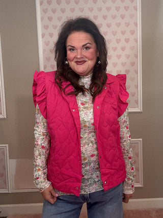 Woman wearing a pink quilted vest over a floral shirt in a room with heart-patterned wallpaper.