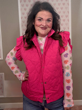 Person wearing a pink quilted vest over a patterned shirt with a heart-themed background.