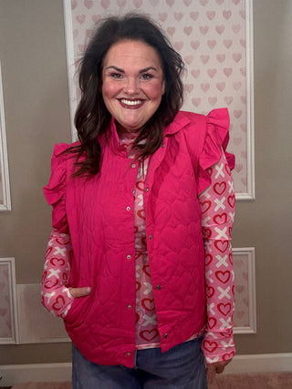Woman wearing a pink quilted vest over a heart-patterned shirt in a room with framed pictures on the wall.