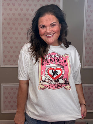 Woman modeling a white Cupid Brewing Co graphic tee featuring a pink vintage love potion design with cupid and heart artwork, styled as casual women’s clothing with relaxed comfy mom style.