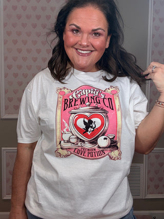 Woman modeling a white Cupid Brewing Co graphic tee featuring a pink vintage love potion design with cupid and heart artwork, styled as casual women’s clothing with relaxed comfy mom style.
