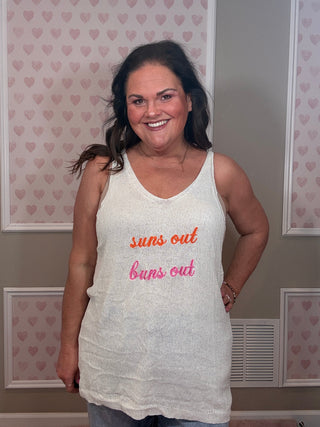 Woman wearing a white tank top with "suns out, buns out" embroidered text, standing in a room with heart-patterned wallpaper.