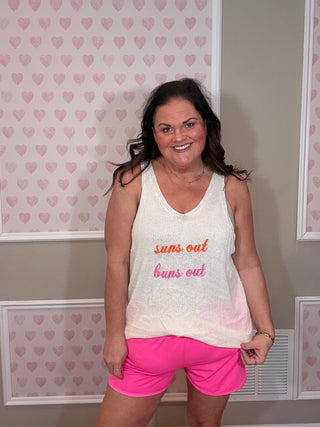 Woman wearing a white tank top with "suns out, buns out" embroidered text and pink shorts in a room with heart-patterned wallpaper.