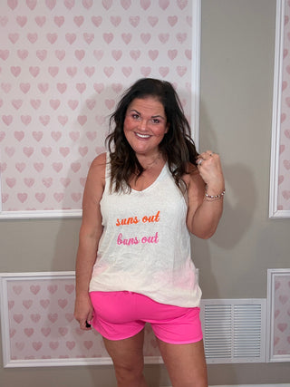 Woman wearing a white tank top with "suns out, buns out" embroidered text and pink shorts in a room with heart-patterned wallpaper.