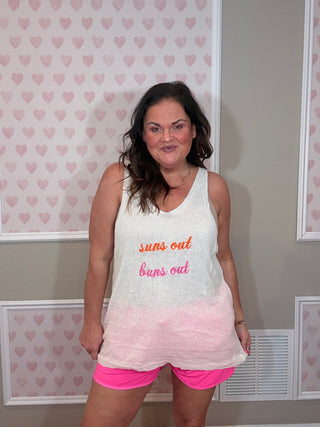 Woman wearing a white tank top with "suns out, buns out" embroidered text and pink shorts in a room with heart-patterned wallpaper.