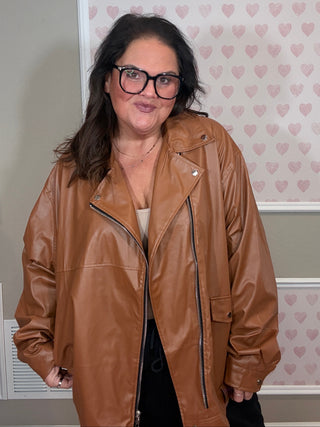 Person wearing an oversized brown leather jacket in front of a wall with heart patterns.