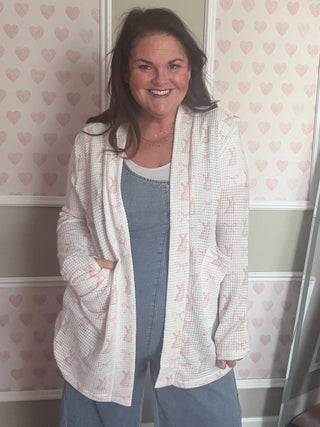 Woman standing in front of a mirror wearing a white cardigan and blue jeans, with a heart-patterned wallpaper background.