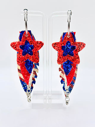Star-shaped earrings with red, white, and blue glittery design on a clear stand against a white background.