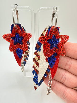 Star-shaped earrings with red, blue, and white glittery design held by a hand.