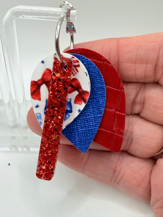Layered patriotic statement earrings with red glitter accent, patriotic bow print, royal blue textured faux leather, red woven texture, and silver hoop closure.