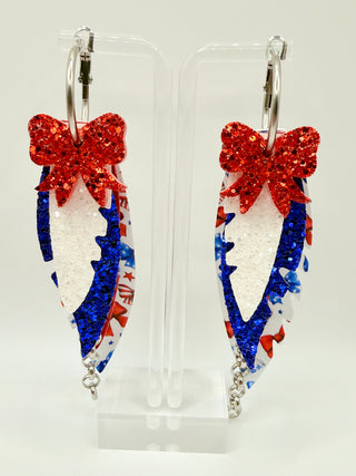 Patriotic bow print statement earrings with red and blue glitter accents, layered faux leather feather details, silver chain charm, and lightweight boutique design.