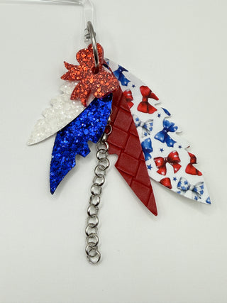 Patriotic bow print statement earrings with red and blue glitter accents, layered faux leather feather details, silver chain charm, and lightweight boutique design.
