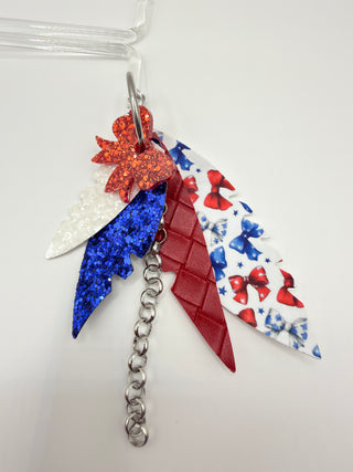 Patriotic bow print statement earrings with red and blue glitter accents, layered faux leather feather details, silver chain charm, and lightweight boutique design.