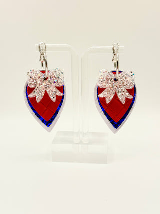 Layered patriotic statement earrings with white glitter bow accent, red woven faux leather texture, royal blue glitter layer, pearl white detail, and silver hoop closure.