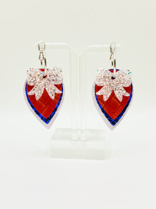 Layered patriotic statement earrings with white glitter bow accent, red woven faux leather texture, royal blue glitter layer, pearl white detail, and silver hoop closure.