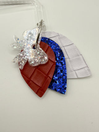Layered patriotic statement earrings with white glitter bow accent, red woven faux leather texture, royal blue glitter layer, pearl white detail, and silver hoop closure.