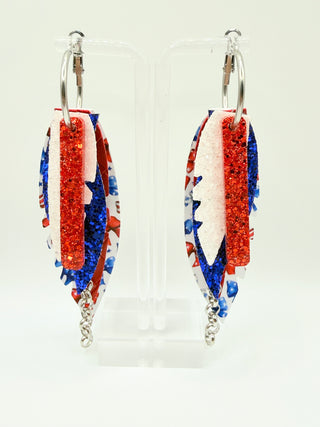 Patriotic bow print statement earrings with red and blue glitter accents, layered faux leather feather details, silver chain charm, and lightweight boutique design.
