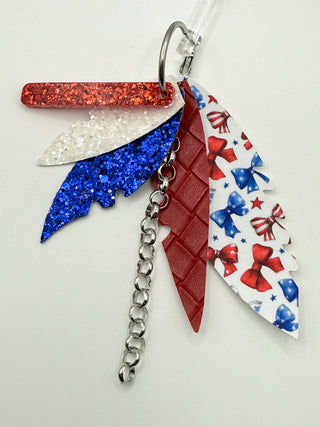 Patriotic bow print statement earrings with red and blue glitter accents, layered faux leather feather details, silver chain charm, and lightweight boutique design.