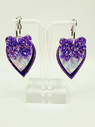 Heart-shaped purple glitter earrings on a clear stand against a white background