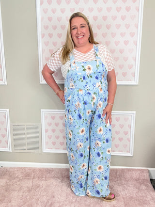 Woman wearing a blue floral overalls standing in a room with framed pictures on the wall.