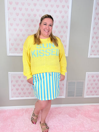Woman wearing a yellow 'SUN KISSED' sweater and striped skirt in a room with heart-patterned wallpaper.