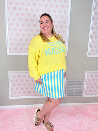 Woman wearing a yellow sweatshirt with 'SUGAR KISSES' text and a striped skirt in a room with heart-patterned wallpaper.