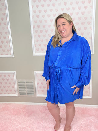 Woman wearing a blue dress standing in a room with heart-patterned wallpaper.