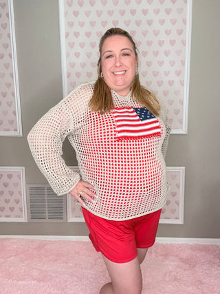 Woman wearing a red and white patterned top with an American flag design, standing in a room with pink walls and framed pictures.