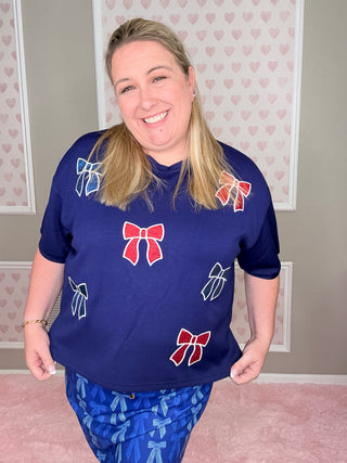 Woman wearing a blue shirt with red and white bow designs, standing against a light-colored wall.