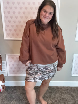 Woman wearing a brown sweatshirt and camouflage skirt standing in a room with heart-patterned wallpaper.