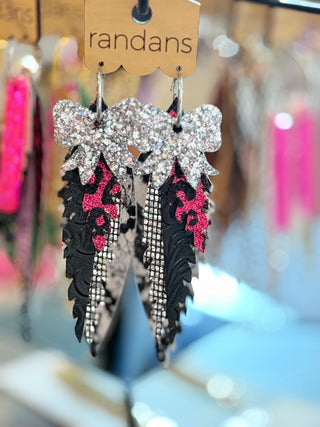 Layered faux leather earrings featuring a sparkly silver bow on top, hot pink glitter cheetah print, black scalloped leather, and rhinestone strips. A bold and glittery statement earring set hanging on a "randans" earring card.