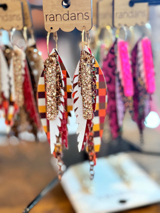 Pair of layered faux leather earrings featuring a fall checkered pattern in orange, yellow, and brown tones with copper glitter accents and white detailing, finished with dangling bronze chains. Displayed on a “randans” card on a rotating earring rack.