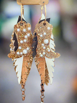 Close-up of glittery gold and bronze layered faux leather earrings with pearl accents, dark brown details, and ivory leather backing, hanging from gold hoops on a “randans” earring card.