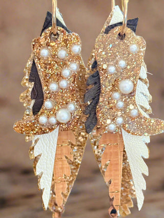 Close-up of glittery gold and bronze layered faux leather earrings with pearl accents, dark brown details, and ivory leather backing, hanging from gold hoops on a “randans” earring card.