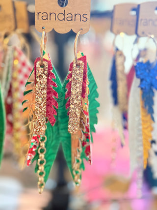 Tiered holiday earrings featuring layers of red glitter, green faux leather, and gold accents with gold hoop hooks and chain fringe, displayed on a "randans" earring card.